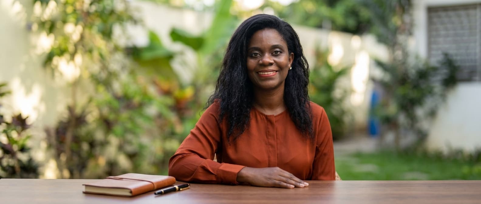 Smiling Black woman in a rust blouse sits at an outdoor table with a notebook.